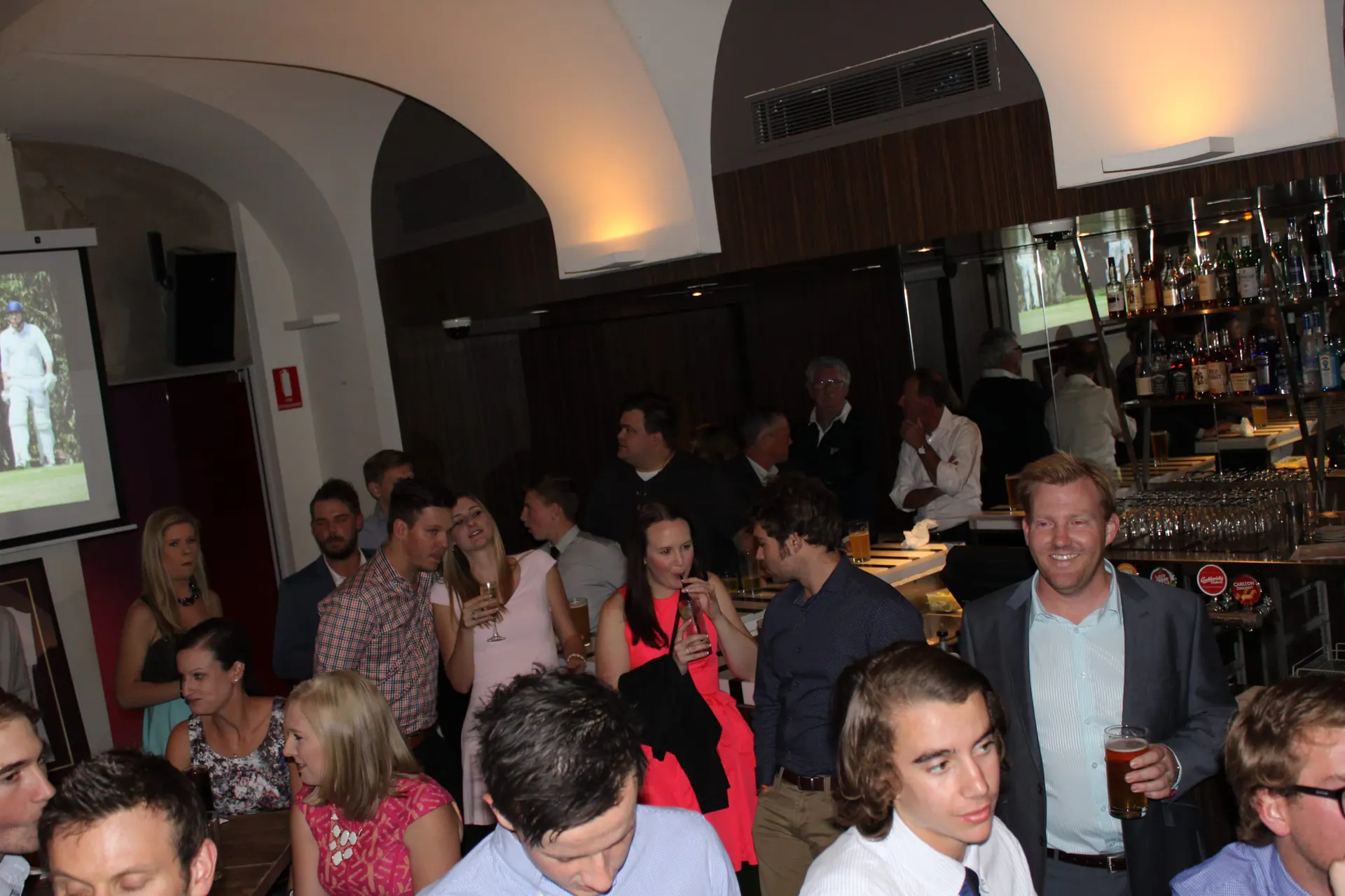 A full function room at a club presentation night, bar at the back and the projector screen running match photos.