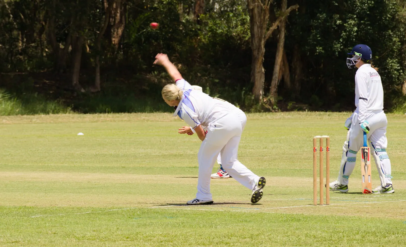 A bowler in whites releases the ball in a women's community match, the batter waiting at the other end.